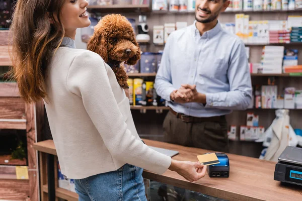 Cropped view of smiling woman holding poodle and paying with credit card near muslim seller in pet shop — Stockfoto