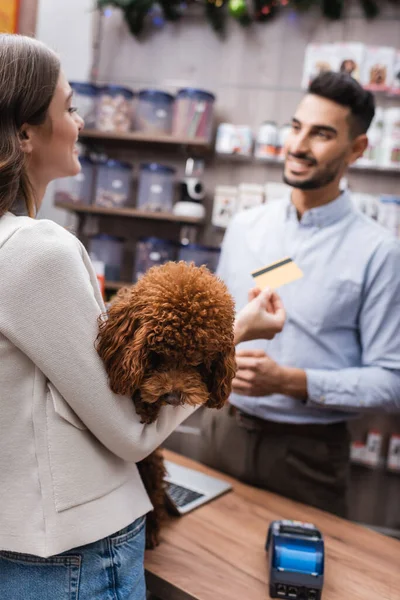 Smiling woman holding poodle and blurred blurred credit card near arabian seller in pet shop — Stockfoto