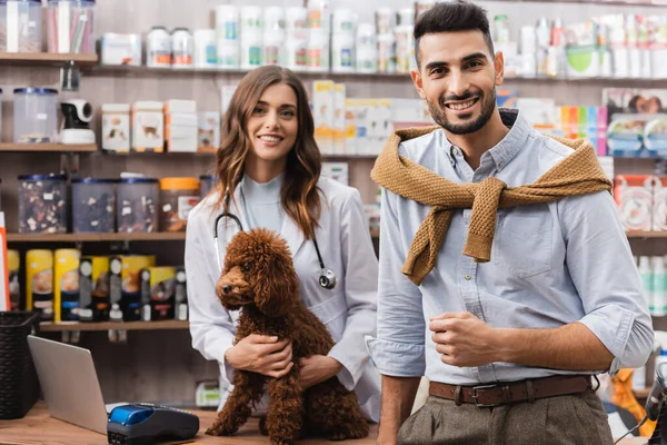 Smiling muslim man looking at camera near veterinarian and poodle in pet shop — Stockfoto
