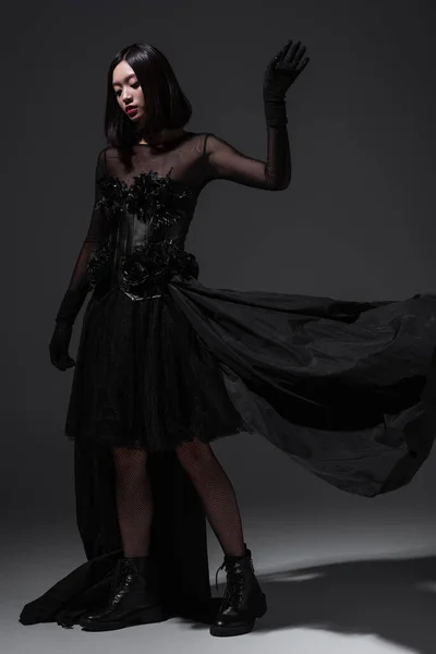 Full length of young asian woman in black gothic dress and gloves posing on dark grey — Stock Photo