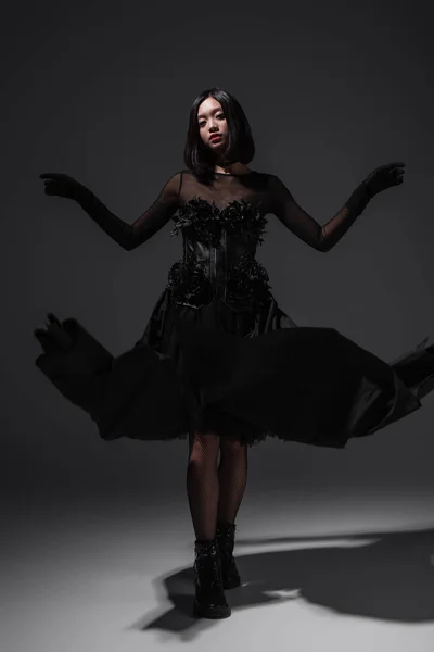Full length of asian young woman in black gothic dress and gloves gesturing on dark grey — Stockfoto