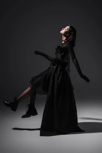 Side view of asian young woman in black gothic dress and boots levitating on dark grey — Stockfoto