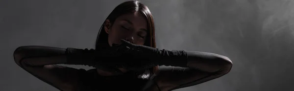 Brunette asian model with closed eyes and gloves posing on dark grey with smoke, banner — Stock Photo