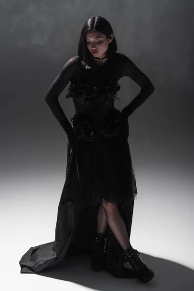 Full length of asian young woman in black gothic dress and gloves posing on dark grey with smoke — Stock Photo