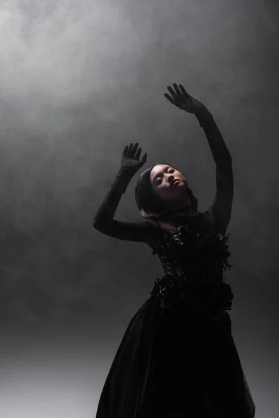 Asian woman with closed eyes in gloves and gothic dress gesturing on grey with smoke — Stock Photo