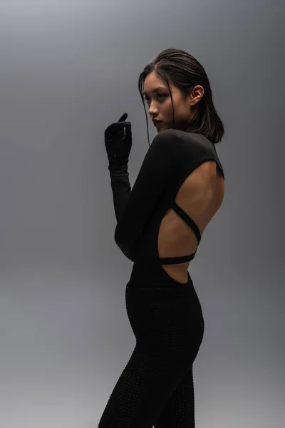 Stylish and young asian model in black outfit with gloves posing isolated on grey — Stockfoto