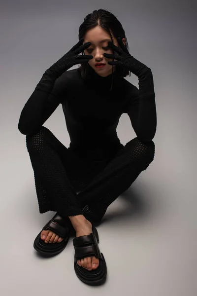 Повно стилізованої азійської жінки в повному чорному одязі і рукавички дотик обличчя на сірому — стокове фото