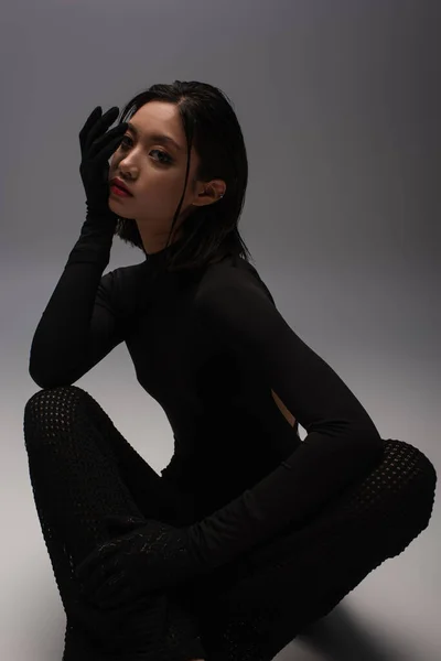 Stylish and young asian model in black outfit with gloves sitting on grey - foto de stock