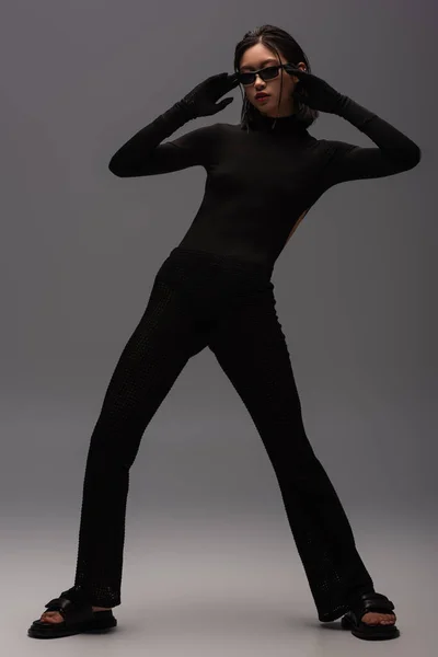 Full length of young asian woman in total black outfit and stylish sunglasses posing on grey — Stock Photo
