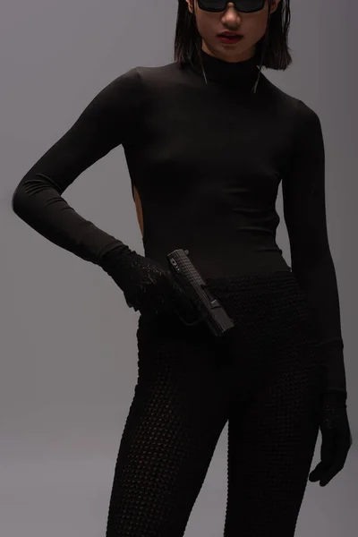 Cropped view of dangerous asian woman in total black outfit and stylish sunglasses holding gun isolated on grey - foto de stock