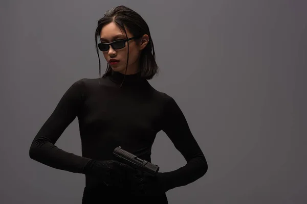 Armed asian woman in total black outfit and stylish sunglasses holding gun isolated on grey - foto de stock