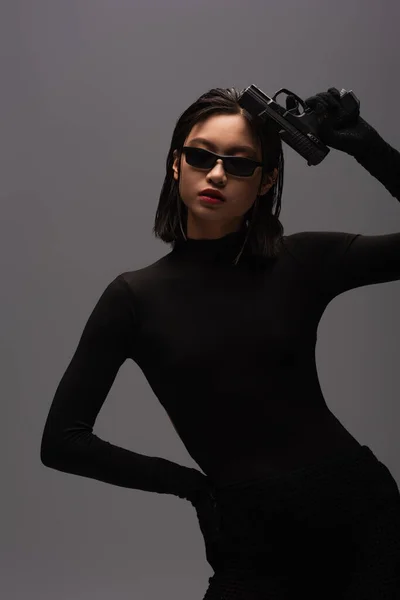 Dangerous asian woman in black outfit and stylish sunglasses holding gun isolated on grey - foto de stock