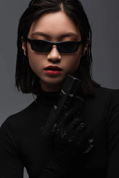 Portrait of dangerous asian woman in black outfit and sunglasses holding gun isolated on grey — Photo de stock