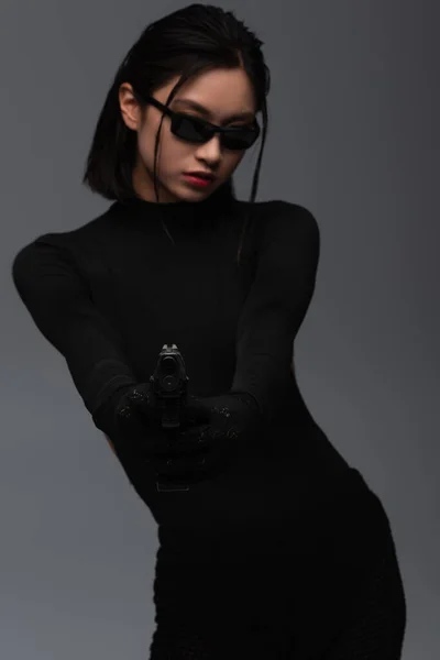 Dangerous asian woman in total black outfit and stylish sunglasses aiming with gun isolated on grey — Photo de stock