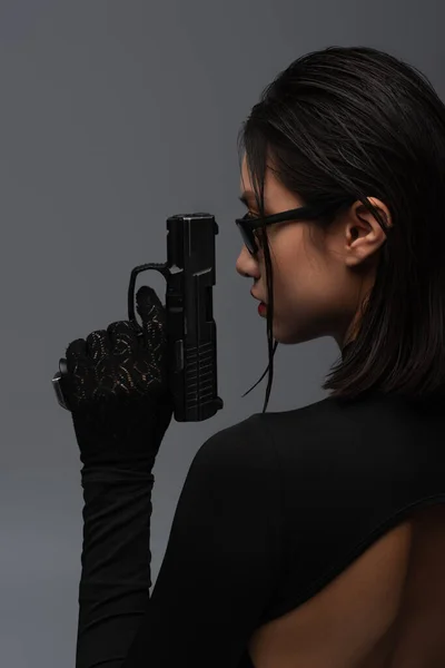 Side view of dangerous asian woman in total black outfit and stylish sunglasses holding gun isolated on grey — Stockfoto