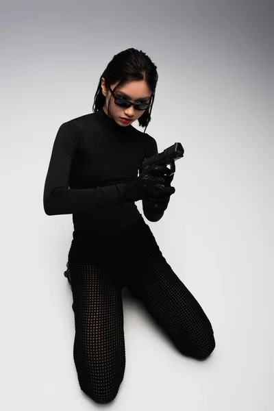 Dangerous asian woman in total black outfit and stylish sunglasses holding gun on white — Photo de stock