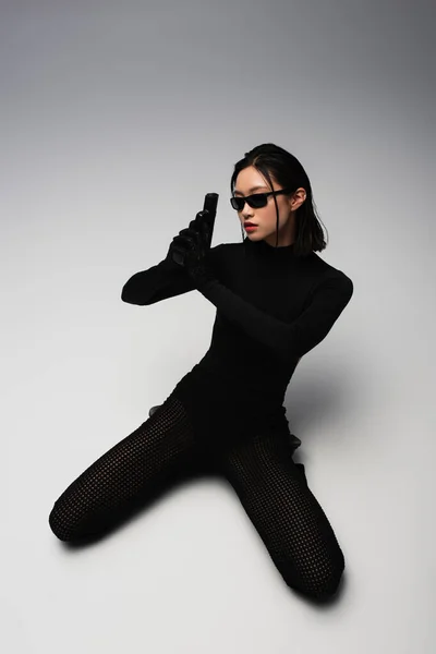 High angle view of dangerous asian woman in total black outfit and stylish sunglasses holding gun on white - foto de stock