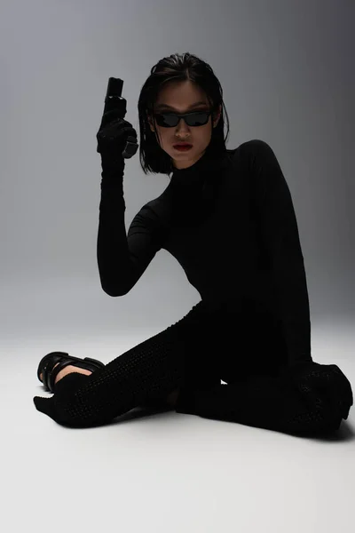 Dangerous asian woman in total black outfit and stylish sunglasses holding gun while sitting on grey — Stockfoto