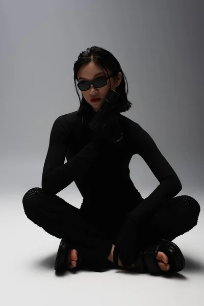 Asian woman in total black outfit and stylish sunglasses holding gun while sitting on grey - foto de stock