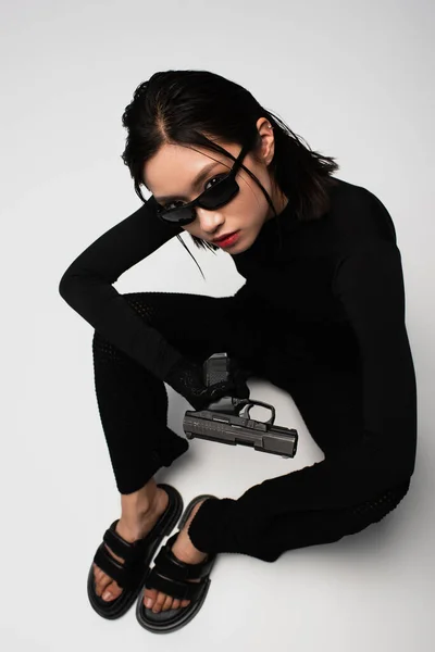 Top view of asian woman in total black outfit and stylish sunglasses sitting near gun on white - foto de stock