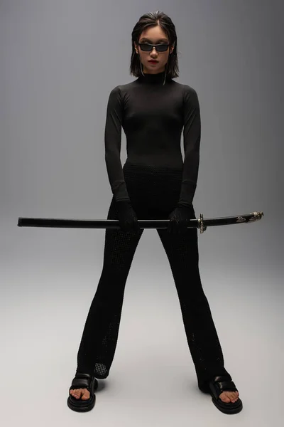 Full length of asian woman in black outfit and stylish sunglasses holding katana sword on grey — Fotografia de Stock