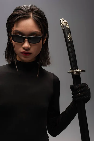 Brunette asian woman in black clothes and stylish sunglasses holding katana sword in scabbard isolated on grey — Fotografia de Stock