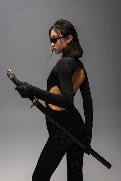 Brunette asian woman in black outfit and stylish sunglasses posing with katana sword isolated on grey - foto de stock