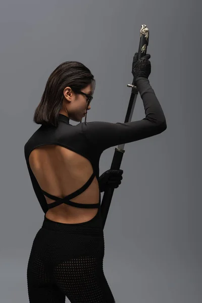 Side view of brunette asian woman in black outfit and stylish sunglasses pulling out katana from scabbard isolated on grey - foto de stock