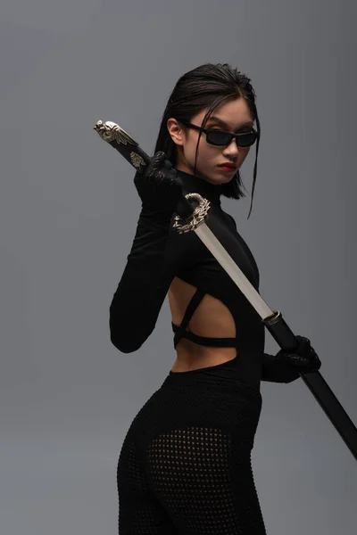 Dangerous asian woman in black outfit and stylish sunglasses pulling out katana from scabbard isolated on grey — Photo de stock