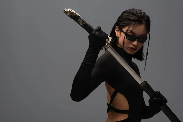 Brunette asian woman in black clothes and sunglasses pulling out katana from scabbard isolated on grey — Photo de stock