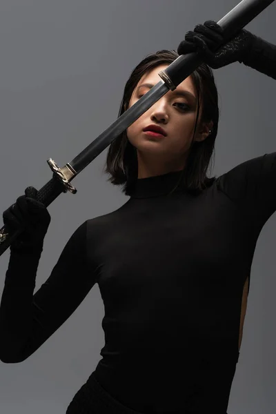 Brunette asian woman in black outfit pulling out katana from scabbard isolated on grey — Stock Photo