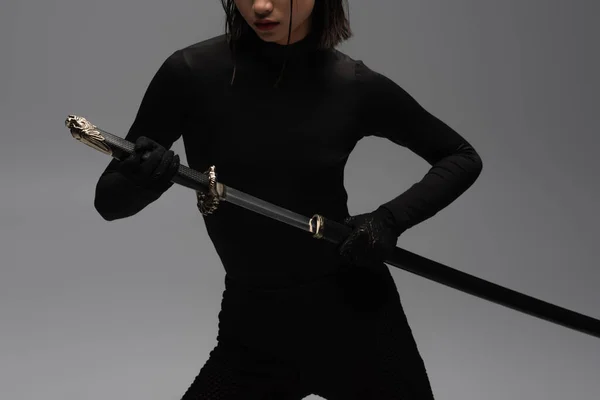 Cropped view of woman in black outfit pulling out katana from scabbard isolated on grey - foto de stock