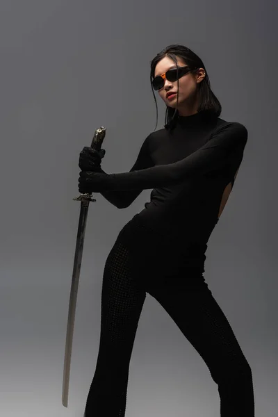 Brunette asian woman in sunglasses and black clothes holding katana isolated on grey - foto de stock