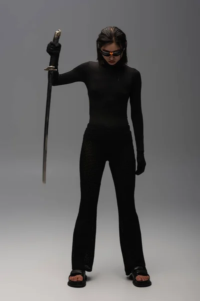 Full length of brunette asian woman in sunglasses and black outfit holding katana on grey — Photo de stock
