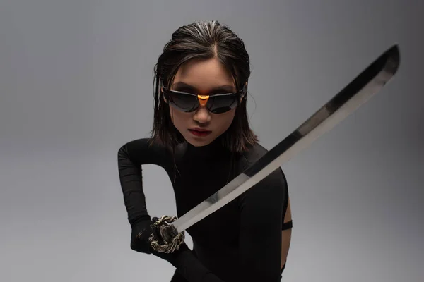 Опасная азиатка в солнцезащитных очках и черном наряде, держащая Катану изолированной на сером — стоковое фото