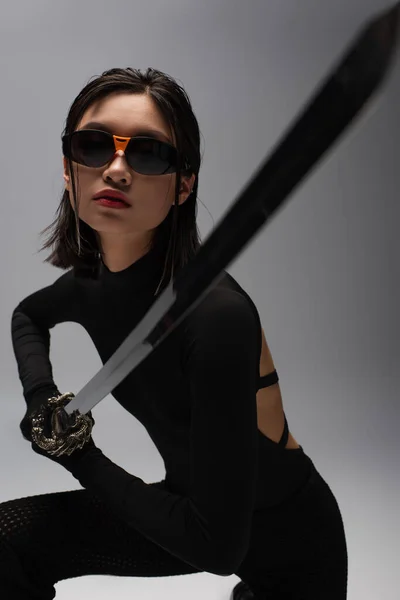 Dangerous asian woman in sunglasses and black outfit holding katana isolated on grey — Fotografia de Stock