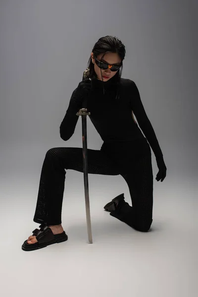 Full length of dangerous asian woman in sunglasses and black clothes holding katana while standing on knee on grey — Photo de stock