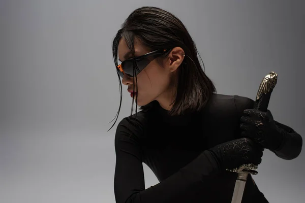 Side view of dangerous asian woman in sunglasses and black outfit holding katana isolated on grey — Fotografia de Stock