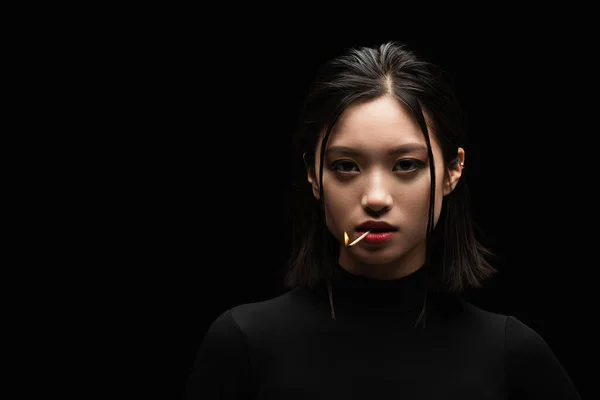 Portrait of pretty asian woman with burning match in mouth looking at camera isolated on black — Stock Photo