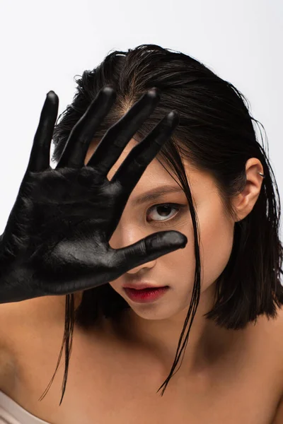 Young asian woman with bare shoulders covering face with hand in black paint isolated on white — Stock Photo