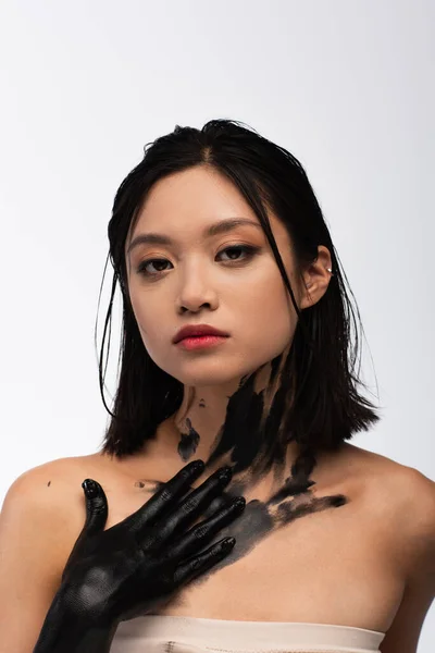 Young asian woman with bare shoulders and hand in black paint touching body while looking at camera isolated on white — Stock Photo
