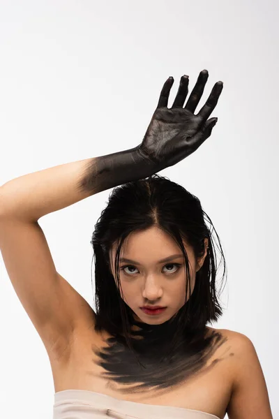 Pretty young asian woman with bare shoulders and hand in black paint above head isolated on white — Stock Photo