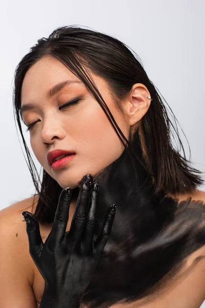 Brunette young asian woman with closed eyes and hand in black paint touching face isolated on white — Stock Photo