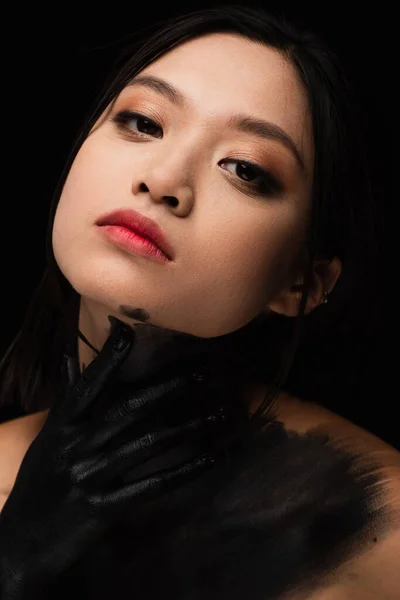 Portrait of young asian woman with hand in paint touching neck isolated on black — Stock Photo