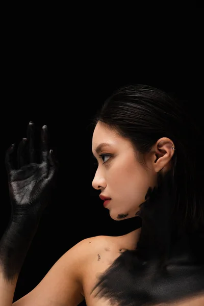 Side view of young asian woman with bare shoulders and hand in paint isolated on black — Stock Photo