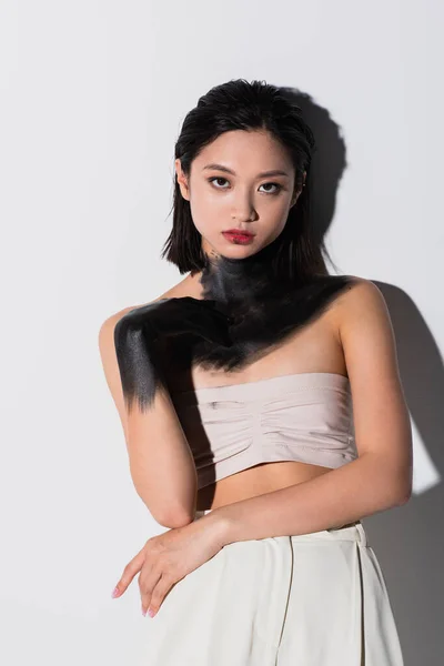 Young asian woman with bare shoulders and hand in black paint looking at camera on white — Foto stock