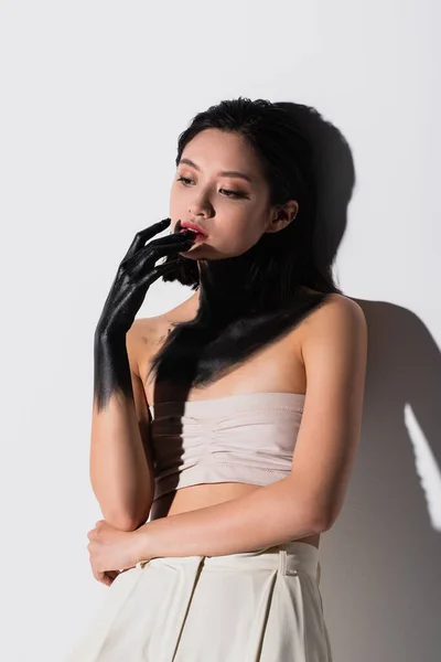 Young asian woman with bare shoulders and hand in black paint touching lip on white — Stock Photo