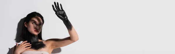 Young asian woman holding hand in black paint above face while covering from light on white, banner — Foto stock