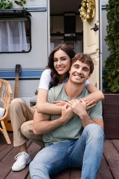 Cheerful couple hugging and looking at camera near blurred camper outdoors — Stock Photo