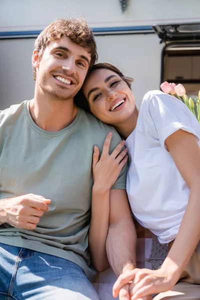 Smiling young couple looking at camera near blurred camper — Stockfoto